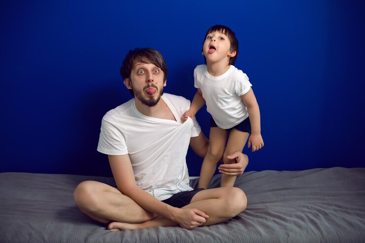 Foto menino com seu pai em camisetas brancas sentado em casa em uma cama contra uma parede azul