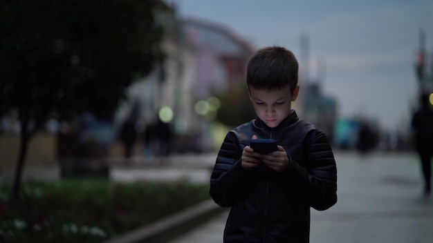 Foto menino com um smartphone moderno caminha pela cidade noturna ao longo da rua