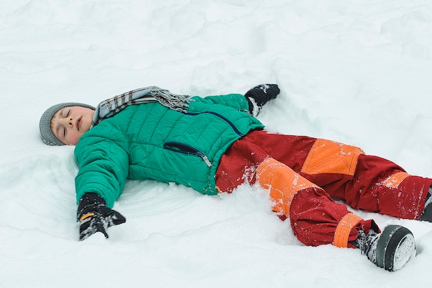 Menino com uma jaqueta verde e calça vermelha deitado de costas na neve com os braços estendidos Dia de inverno