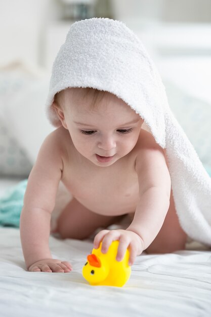 Menino com uma toalha com capuz brincando com um pato de borracha amarela na cama depois do banho