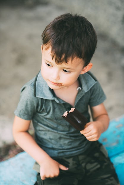 Menino comendo sorvete em um palito coberto de chocolate, fofo ao ar livre