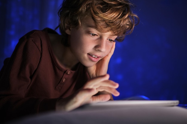 Menino concentrado com cabelo encaracolado em pijama navegando na internet e olhando para a tela enquanto descansa no quarto