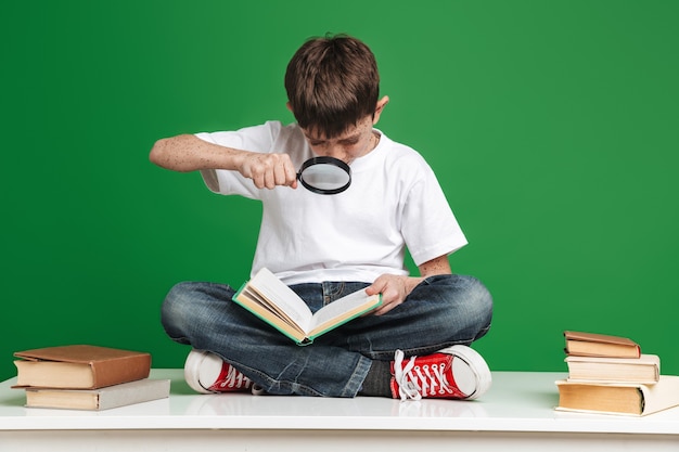 Menino concentrado lendo livro com lupa enquanto está sentado na mesa sobre a parede verde