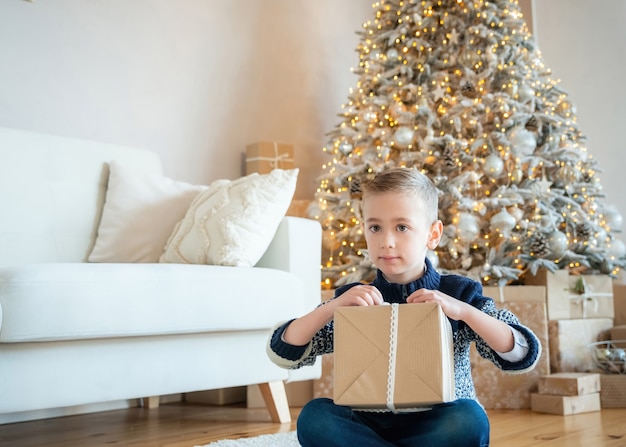 Menino criança desempacotando o presente de Natal. Garoto de cabelo loiro bonito perto da árvore de Natal com caixa de presente. Sala de estar decorada. Tempo mágico. Véspera de Ano Novo.