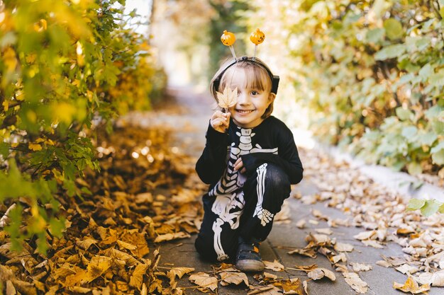 Foto menino criança em fantasia de carnaval de esqueleto de halloween sentado em folhas secas no parque no outono dia de todos os santos férias de outono da escola