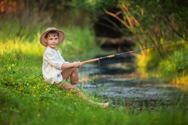 menino criança em roupas rústicas e um chapéu de vime senta-se com uma vara de pescar nas mãos e pega peixe na margem do rio. hobbies e lazer para homens