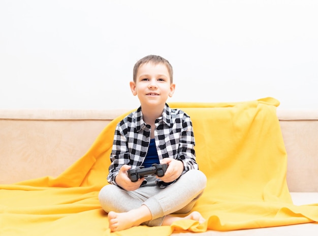 Menino criança feliz na camisa sentada no sofá com joystick preto nas mãos jogando o videogame Jogando videogame no conceito de casa