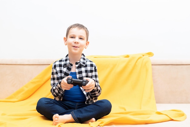 Menino criança feliz na camisa sentada no sofá com joystick preto nas mãos jogando o videogame Jogando videogame no conceito de casa