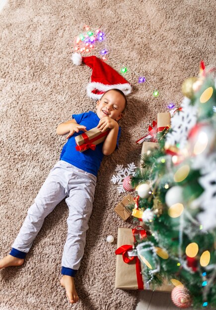 Menino criança feliz na manhã de natal recebe presente deitado no tapete aconchegante em seu quarto de pijama