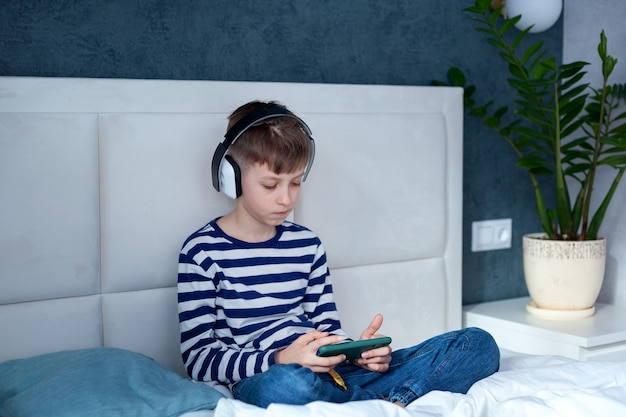 Menino criança no fone de ouvido olhando e jogando telefone, aprendendo na cama na sala de estar. Crianças do conceito usando tecnologia