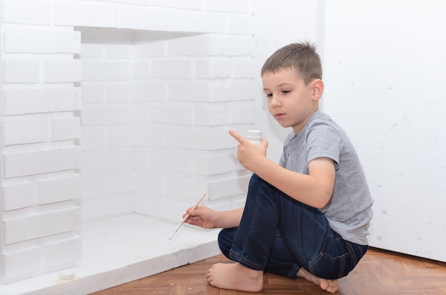 Menino criança senta-se no chão e pinta uma lareira decorativa com guache de tinta branca Melhoria da casa conceito DIY Foco seletivo