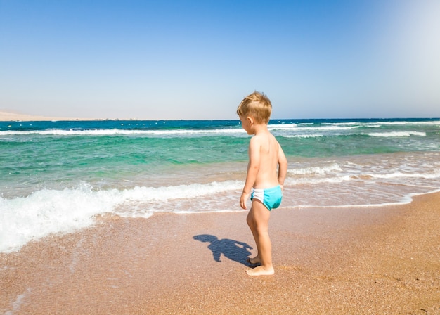 Menino da criança andando na praia e olhando as ondas do mar calmo e quente. Criança relaxando e se divertindo durante as férias de verão.