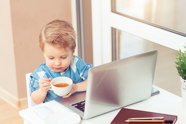 Menino da criança bebendo chá e fazendo anotações, usando o laptop. Conceito de negócio de bebê