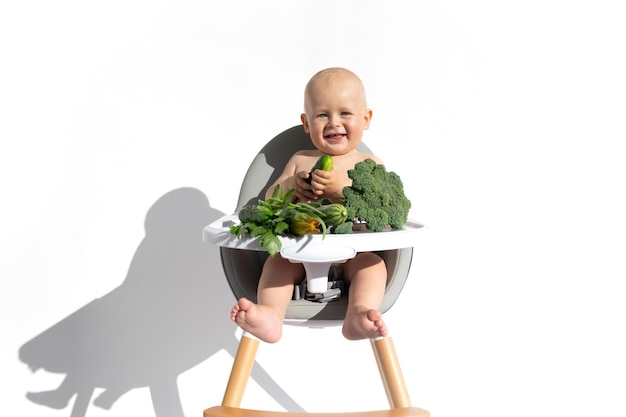 Menino da criança na cadeira de alimentação come vegetais orgânicos salada pepinos cebola brócolis Sombra dura isolada no fundo branco Dieta saudável vegana alimentando o conceito de comida sólida Copiar espaço