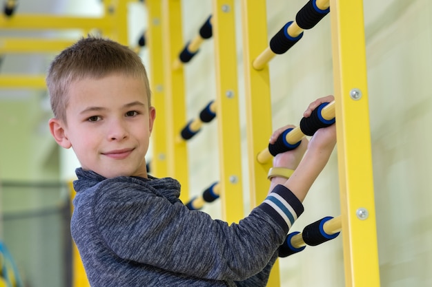 Menino da jovem criança que exercita em uma barra da escada da parede dentro da sala do gym dos esportes em uma escola.