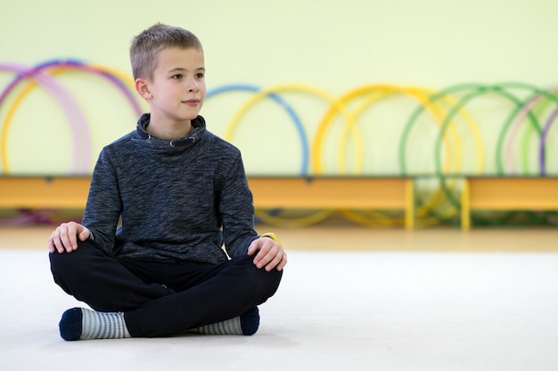 Menino da jovem criança que senta-se e que relaxa no assoalho dentro da sala de esportes em uma escola após o treinamento.