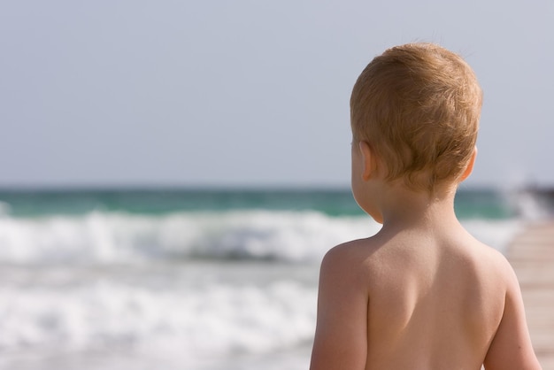Menino de 2 anos na praia de areia Pronto para nadar Olhando para as ondas da costa