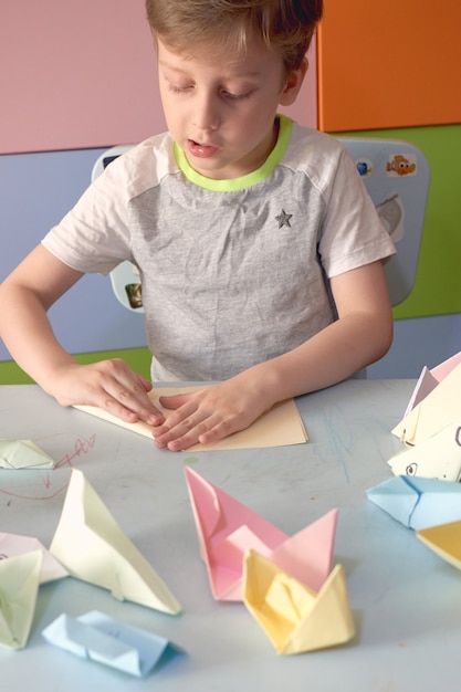 Menino de 6 anos faz um origami de aviões e sapos durante a quarentena Covid-19, auto-isolamento, conceito de educação online, ensino doméstico. Criança em casa, jardim de infância fechado, arte infantil.