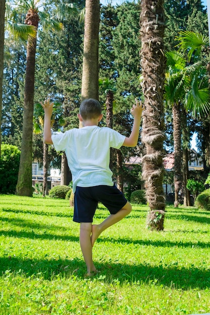 Menino de 7 anos puxa as mãos para o sol fazendo ioga e olha para a vista traseira das palmeiras Sonhando e meditando zen como conceito Relaxamento viagens férias crianças