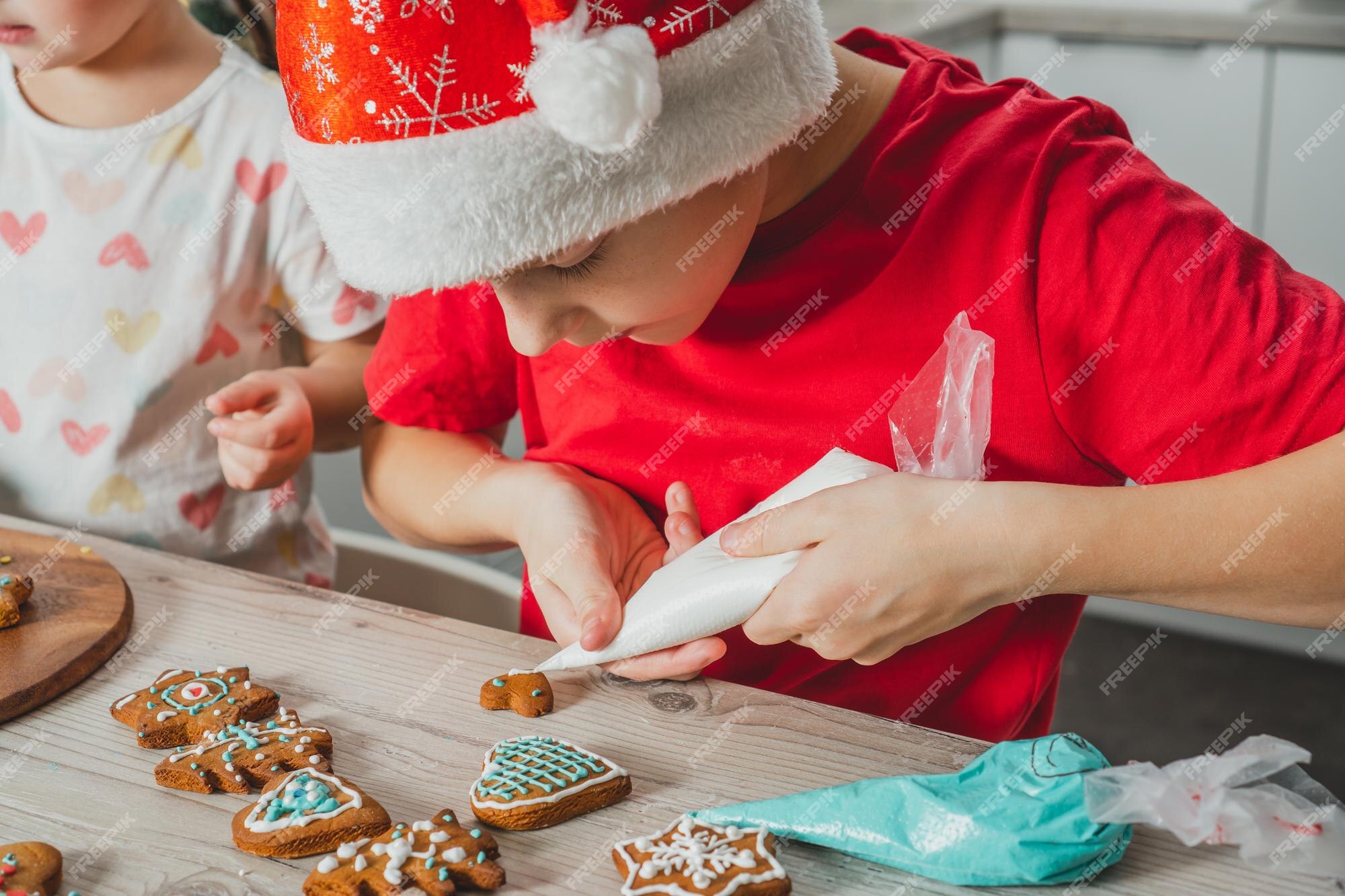 Menino de 8 anos com chapéu de papai noel vermelho decora biscoitos de gengibre de natal com cobertura na mesa de madeira da cozinha | Foto Premium Menino de 8 anos com chapéu de papai noel vermelho decora biscoitos de gengibre de natal com cobertura na mesa de madeira da cozinha | Foto Premium