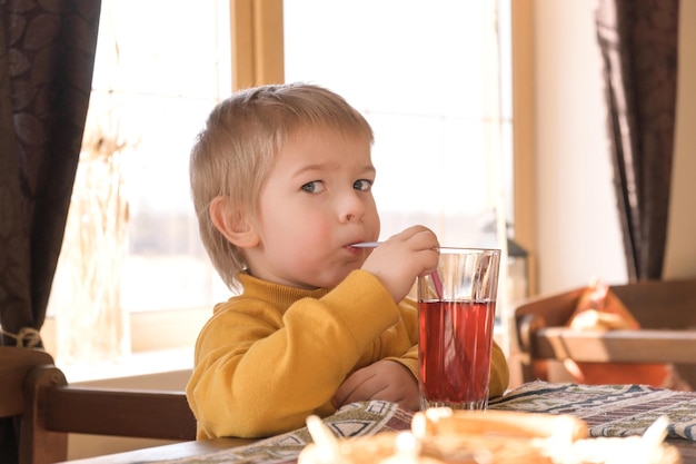 Menino de cabelo loiro bebendo limonada no café, menino de 3 anos bebe suco vermelho de vidro