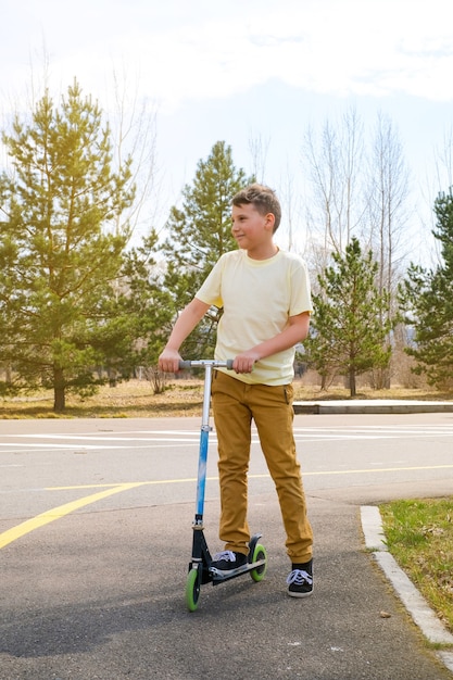 Menino de calça amarela na estrada do parque com scooter elétrica Dia de sol de primavera