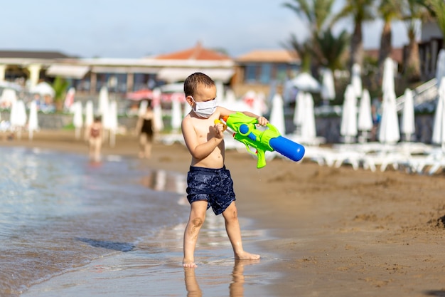 Menino de calção de banho e arma de brinquedo com máscara médica na praia se protegendo do vírus corona