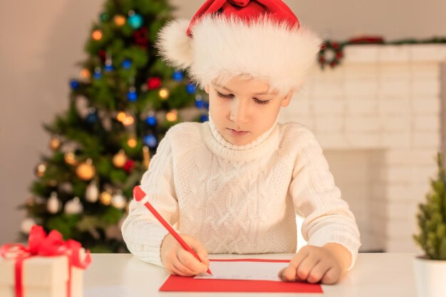 Menino de chapéu de papai noel vermelho escrevendo uma carta para o natal de papai noel ou feriados aconchegantes de ano novo