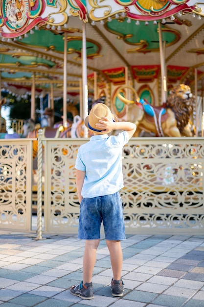 Menino de chapéu em um parque de diversões fica na frente de um carrossel de férias