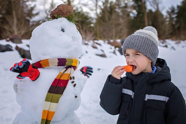 Menino de chapéu morde cenoura que era o nariz de boneco de neve sorrindo estilo de vida de fim de semana familiar de inverno