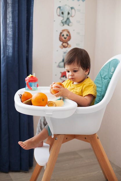 Foto menino de criança com cabelo escuro em uma camiseta amarela, senta-se em uma cadeira alta e janta frutas.