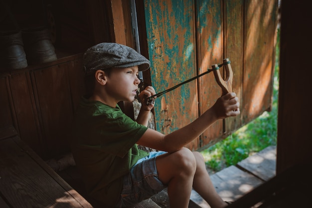 Foto menino de criança em um boné mira com um estilingue para atirar em um alvo. brincar como uma criança na aldeia de férias.