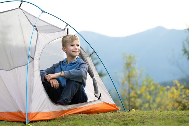 Menino de criança feliz descansando em uma barraca de turista no acampamento de montanha, apreciando a vista da bela natureza de verão. Caminhadas e conceito de modo de vida ativo.