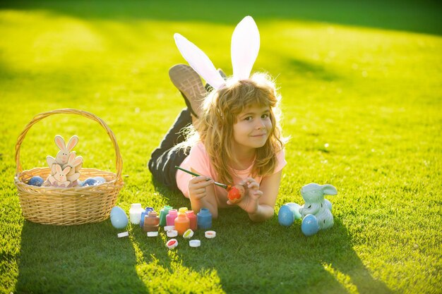 Menino de crianças de páscoa em orelhas de coelho pintam ovos de páscoa criança fofa ao ar livre em fantasia de coelho com orelhas de coelho...