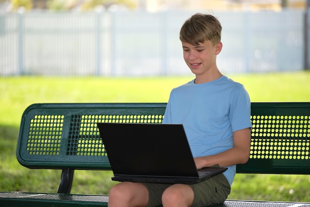 Menino de escola adolescente digitando no computador portátil sentado no banco ao ar livre no dia ensolarado de verão Educação durante o conceito de quarentena