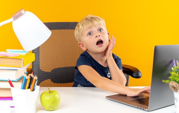 Menino de escola assustado sentado à mesa com as ferramentas da escola usando o laptop colocando a mão na bochecha