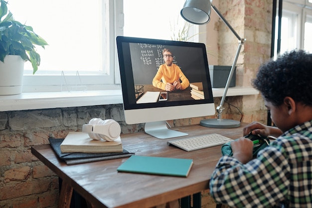 Menino de escola contemporâneo sentado à mesa em frente ao monitor do computador durante a aula on-line e tirando uma caneta ou lápis da caixa de lápis