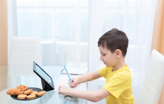 Menino de escola em camiseta amarela sentado à mesa com tablet digital estudando em casa Conceito de educação on-line de ensino à distância