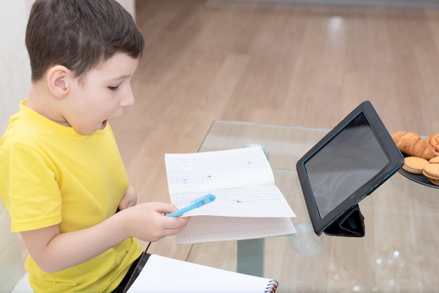 Menino de escola em camiseta amarela sentado à mesa com tablet digital estudando em casa Conceito de educação on-line de ensino à distância