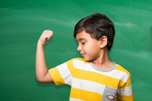 Menino de escola indiano fofo na mão esticada pose sobre lousa verde ou fundo de quadro de giz segurando livros, copo da vitória etc, isolado