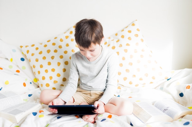 Menino de escola na cama em casa com tablet digital pad na mão, fazendo lição de casa. Educação online a distância. Quarentena. Jogos. Menino brinca com smartphone