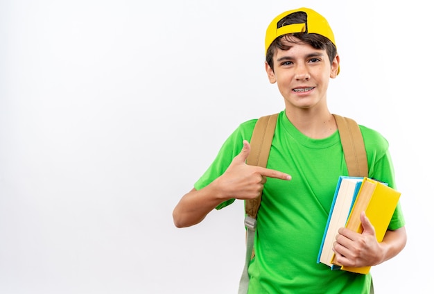 Menino de escola satisfeito usando uma mochila com um boné segurando e apontando para os livros