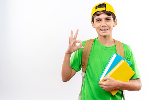 Menino de escola sorridente usando mochila com tampa mostrando um gesto de aprovação segurando livros