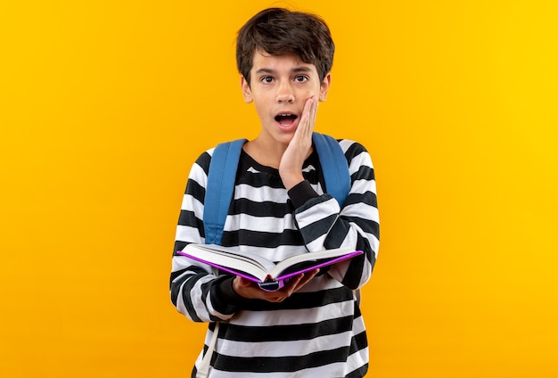 Menino de escola surpreso usando uma mochila segurando um livro e colocando a mão na bochecha isolada em uma parede laranja