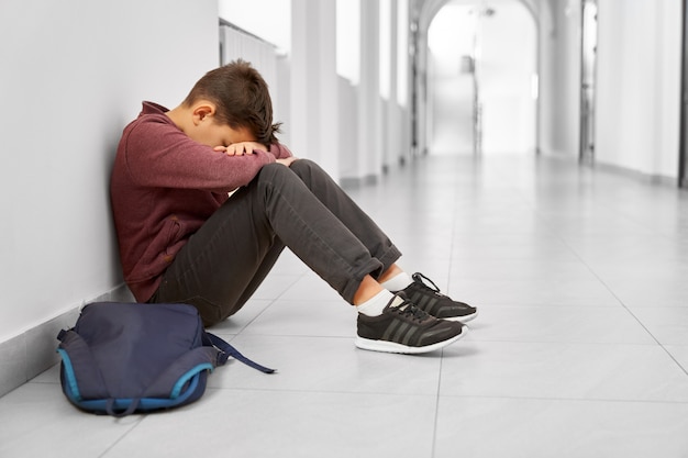 Foto menino de escola triste sentado sozinho no chão no corredor.