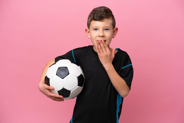 Menino de jogador de futebol caucasiano isolado em fundo rosa feliz e sorridente cobrindo a boca com a mão