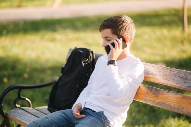 Menino de moletom branco com mochila preta sentado no banco do parque e falar com alguém pelo telefone