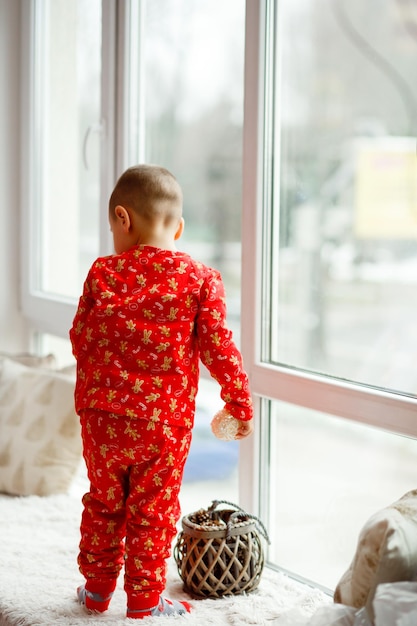 Menino de Natal de pijama vermelho na janela olha para o Papai Noel para receber presentes para o feriado