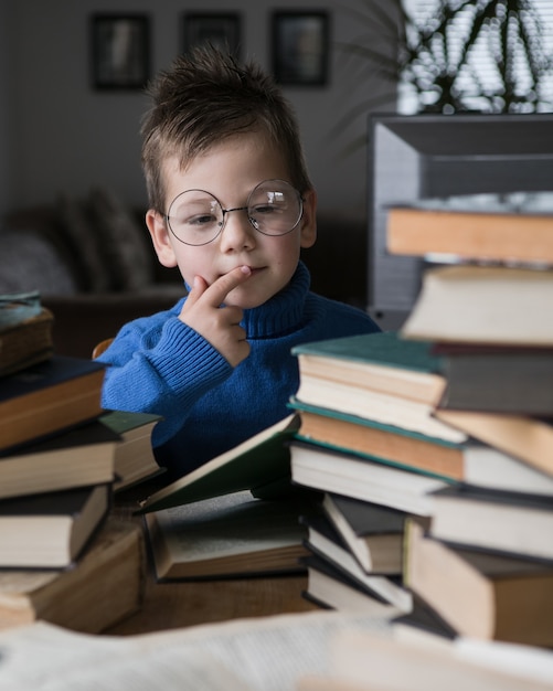 Menino de óculos lendo um livro com uma pilha de livros ao lado dele.