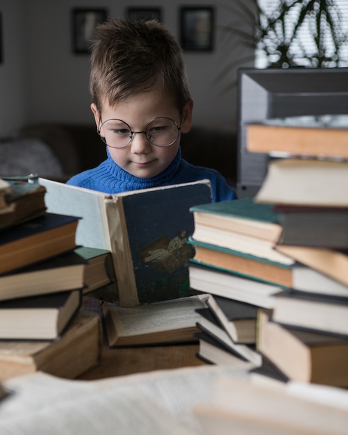 Menino de óculos lendo um livro com uma pilha de livros ao lado dele.